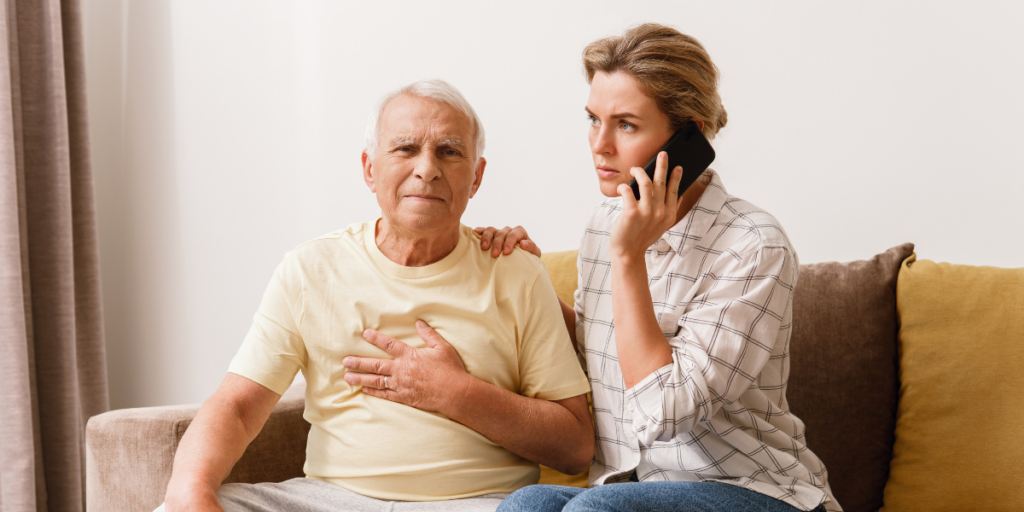 An elderly man clutching his chest while a concerned woman calls for help on her phone.