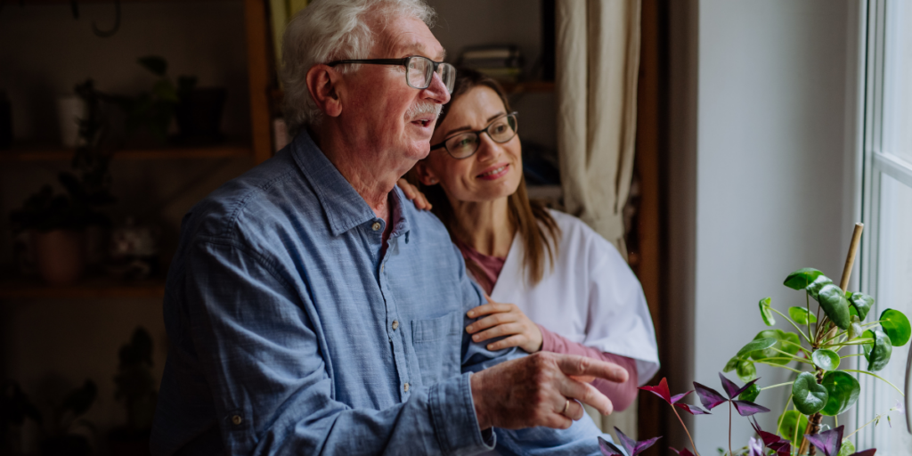 An elderly man and a young woman smile while looking out a window, surrounded by houseplants.