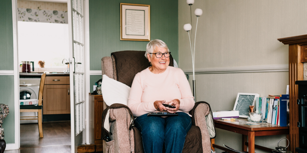 2024_KVY_AgingInPlaceBlogImage2 Smiling older woman sitting in an armchair, holding a remote control in a cozy living room.