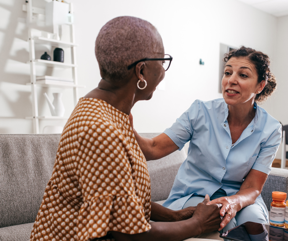 Post 4-nologo A nurse comforts an older woman on a couch, holding her hand and smiling in a bright room.