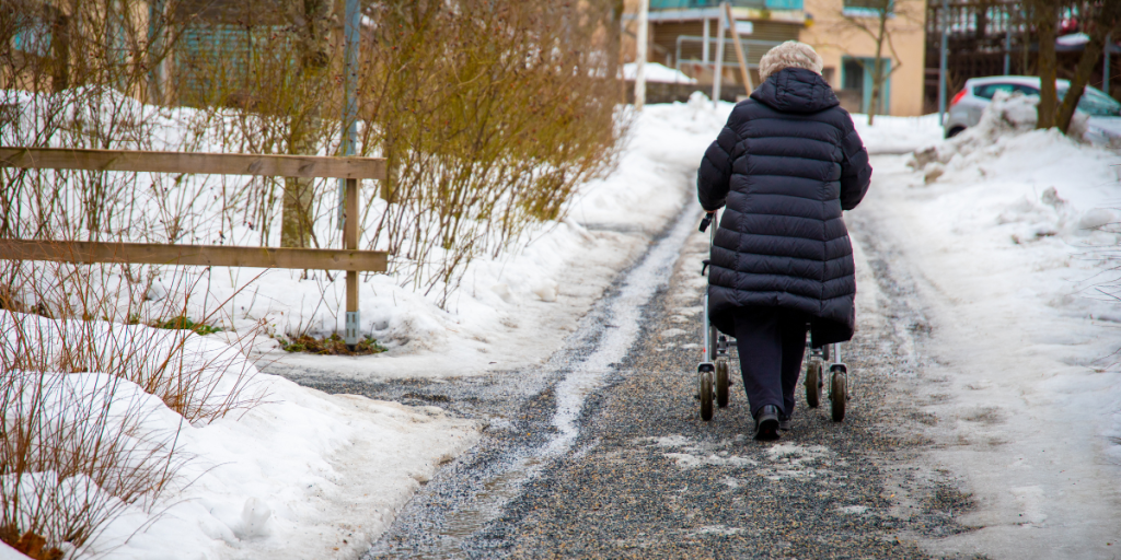 20241216_KVY_HolidaySeasonSafetyBlogSubImage2 Older adult in a black coat walks on a snowy path using a walker, seen from behind.