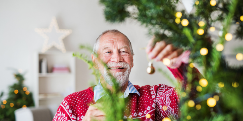 Smiling older man in a festive sweater decorates a Christmas tree with ornaments and lights.