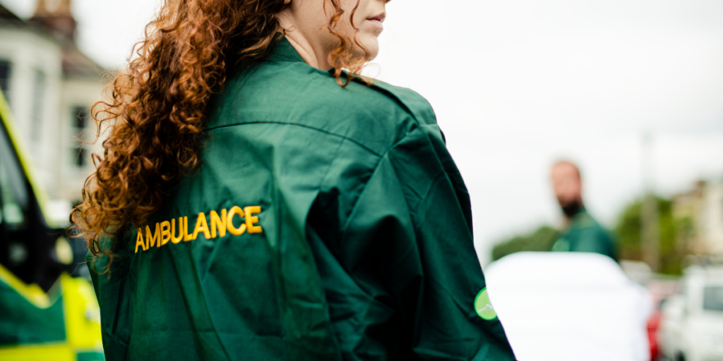 Paramedic with curly hair wearing a green "Ambulance" jacket stands outdoors near an ambulance.