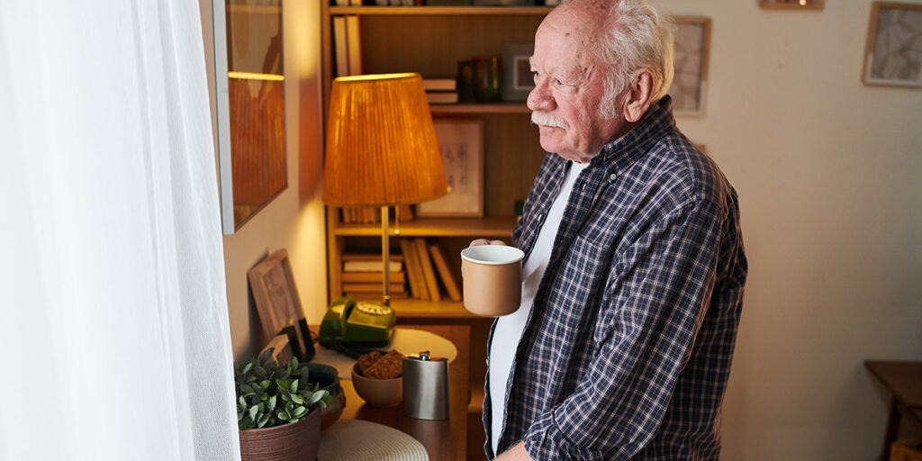 An elderly man holding a mug looks out a window in a cozy, warmly lit room with bookshelves.