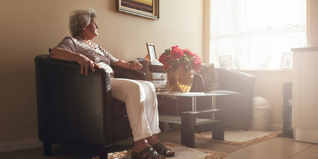 An elderly woman sits alone in an armchair, gazing out a sunlit window in a cozy living room.