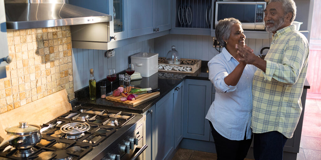 Older couple dancing together and smiling in a cozy kitchen with food and cookware on the counter.