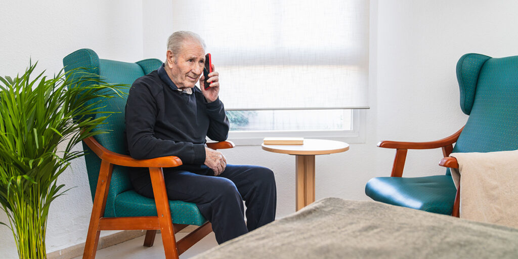 An elderly man sits in a chair, smiling while talking on a mobile phone in a bright, cozy room.