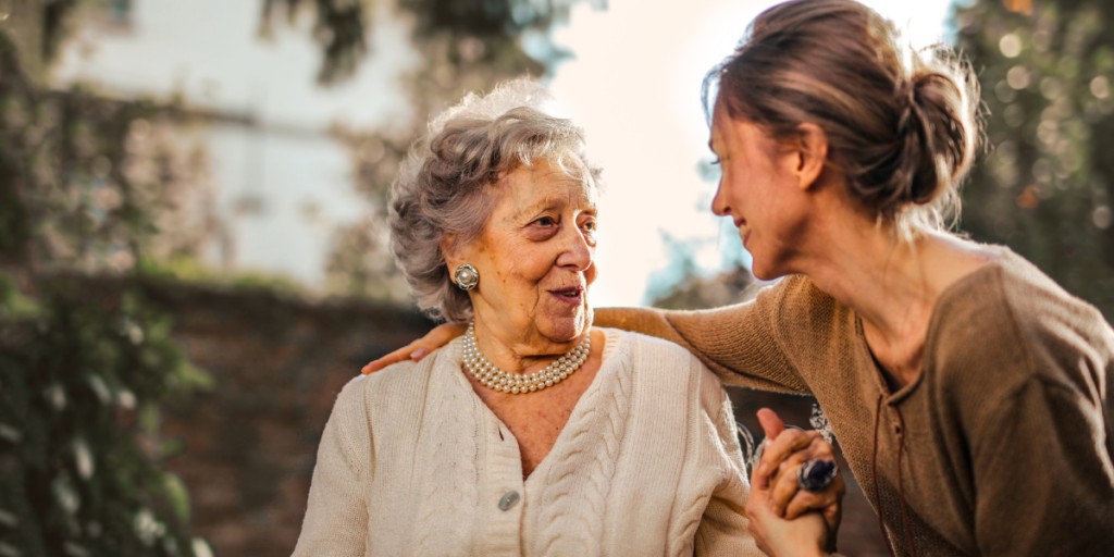 An elderly woman smiles at a younger woman outdoors as they hold hands and share a warm moment.