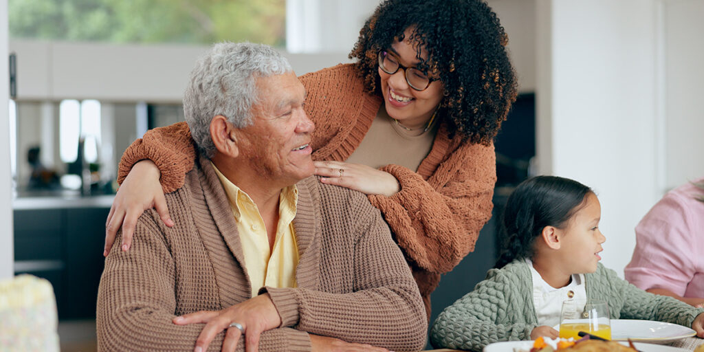 Smiling woman hugs an older man at a table, while a young girl sits beside them, all wearing sweaters.