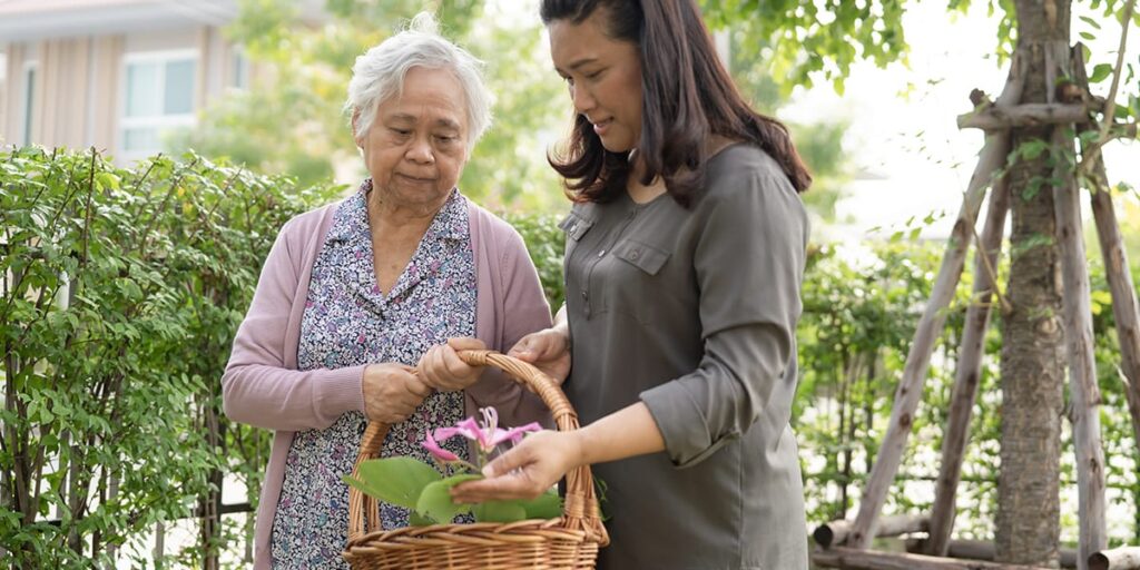 An elderly woman and a younger woman look at flowers in a wicker basket outdoors near greenery.