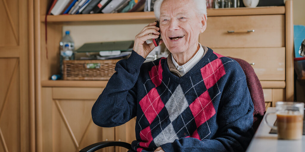 Smiling elderly man in an argyle sweater talking on the phone while sitting at a desk in a cozy room.