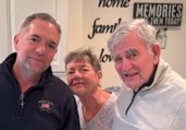 Three adults, two men and one woman, smiling and posing together indoors near a wall with decorative words.