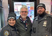 Three smiling people, two in NYPD uniforms, pose together indoors near a glass door.