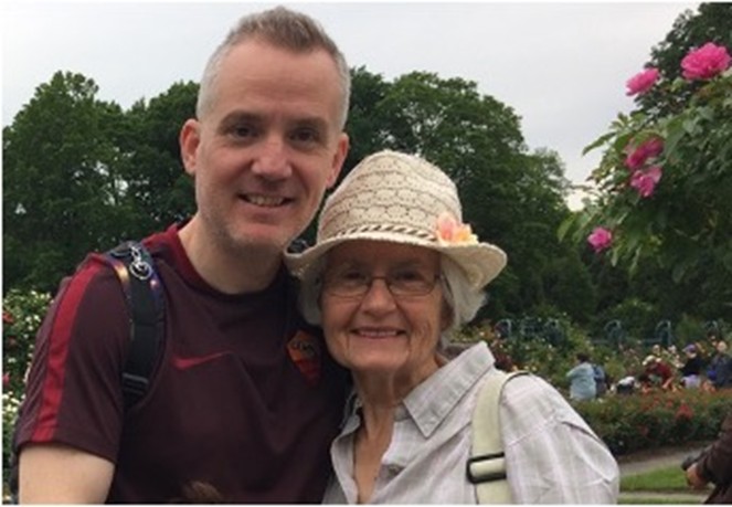 A smiling man and elderly woman in a hat pose together outdoors near blooming flowers and greenery.