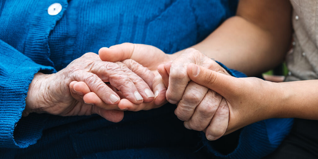 Young hands gently hold the hands of an elderly person wearing a blue sweater, showing care and support.