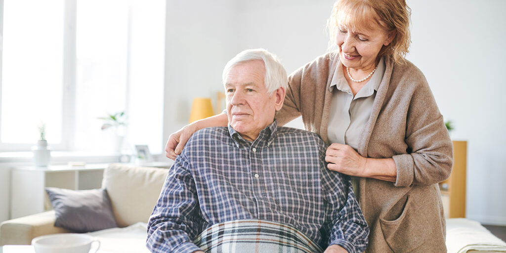 An older woman stands beside and comforts an older man sitting on a couch with a blanket on his lap.