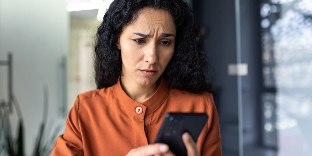 Woman with curly hair in an orange shirt looks worried while reading something on her smartphone.