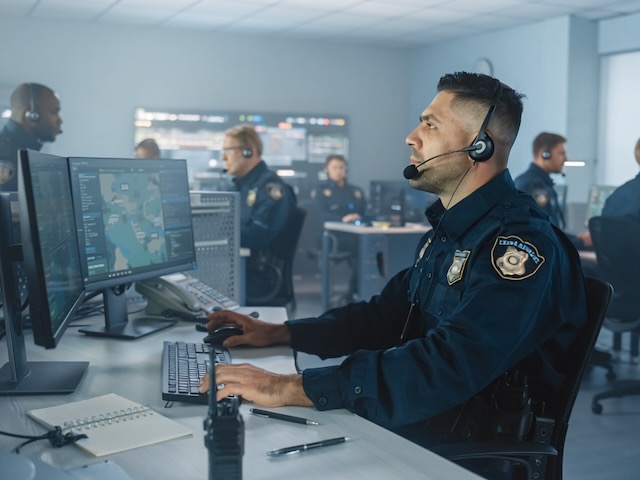 Police officers in a control room monitor screens and communicate using headsets at their desks.