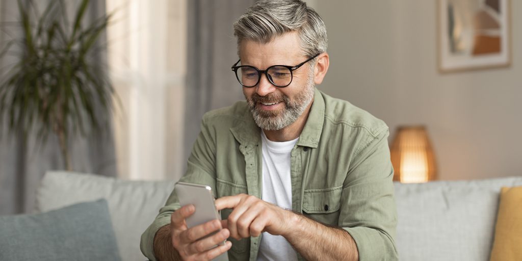 Smiling man with glasses sits on a couch, using a smartphone in a cozy living room.