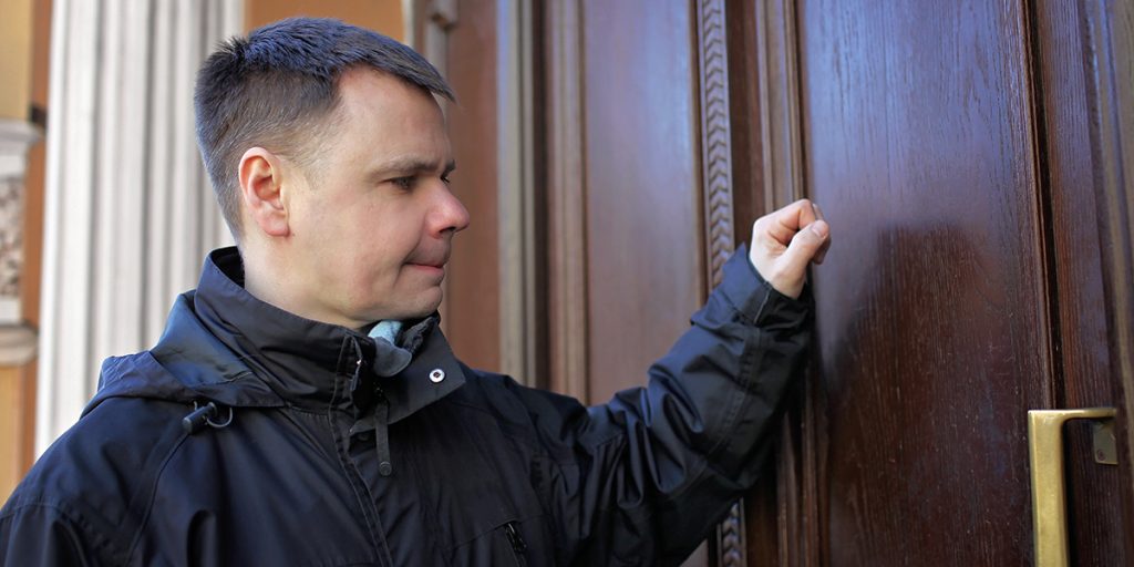 Man in a black jacket knocking on a large brown wooden door with a brass handle.